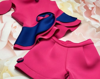 May include: A pink and blue two-piece outfit for a child. The top has a ruffled skirt with a blue trim and a pink rose embellishment. The shorts are solid pink with a pink rose embellishment.