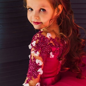 May include: A young girl wearing a pink dress with a burgundy lace bodice and white floral embellishments. Her hair is styled in loose curls and she is looking over her shoulder.