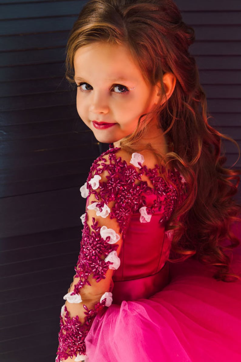 May include: A young girl wearing a pink dress with a burgundy lace bodice and white floral embellishments. Her hair is styled in loose curls and she is looking over her shoulder.