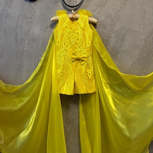 May include: A yellow sleeveless dress with a cape. The dress has a floral pattern and a bow at the waist. The cape is made of sheer fabric.
