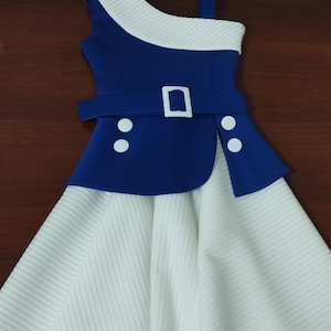 Op de afbeelding: Een blauw en witte jurk met een uniek ontwerp. De jurk heeft een asymmetrische halslijn, een witte riem met een vierkante gesp en een witte rok met verticale strepen. Twee witte knopen zitten aan de voorkant.