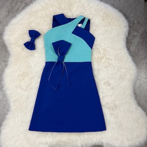 Op de afbeelding: Een kleurgeblokte jurk in blauw- en tinten, met een grote strik in de taille en een bijpassende haaraccessoire. De jurk heeft een asymmetrische halslijn en een eenvoudige A-lijn silhouet.