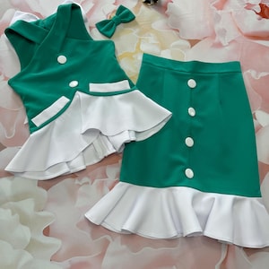 Op de afbeelding: Een groene en witte rok en vest set met een witte strik. De rok en het vest hebben een witte ruche rand. Het vest heeft twee witte knopen en twee zakken.