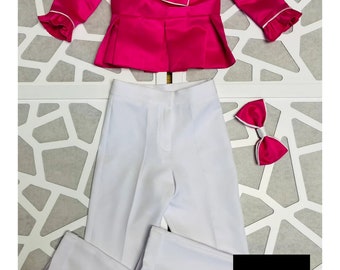 May include: A pink satin blouse with a white trim and a large bow at the neckline. White pants with a cuffed hem. A pink bow tie. The text "Quite the Dress" is printed on a black square.