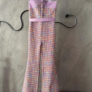 May include: A lavender tweed jumpsuit with a cross-strap bodice and flared legs. The jumpsuit features a matching belt and is adorned with colorful flecks. It hangs on a wooden hanger against a gray background.