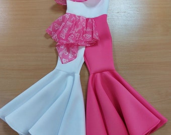 Puede incluir: Un mono para niña blanco y rosa con una superposición de volantes de gasa transparente con la palabra "Barbie" impresa en blanco. El mono tiene una pierna acampanada y un corpiño blanco con un volante rosa.
