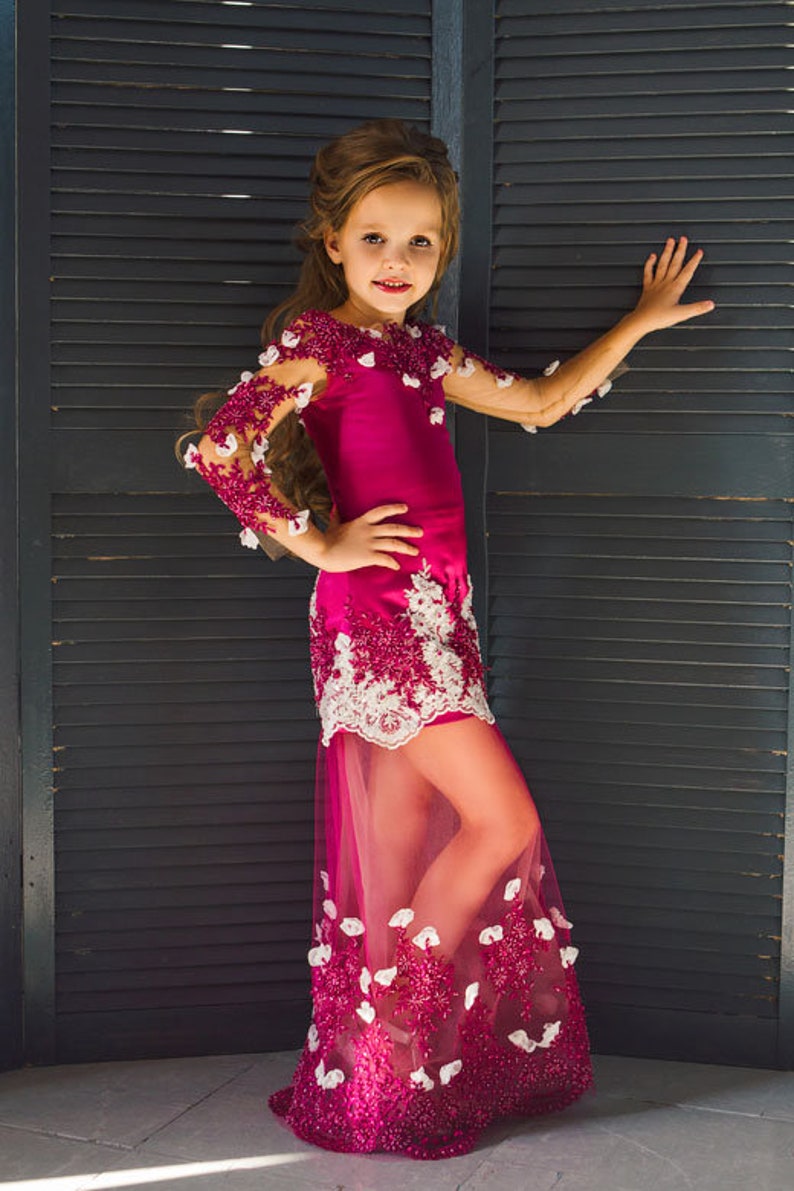 May include: A young girl wearing a pink and white dress with a tulle skirt and floral embellishments. The dress has a fitted bodice and long sleeves. The girl is standing in front of a dark wooden slatted wall.