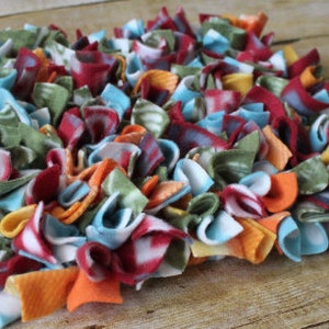 May include: A colorful, handmade fleece dog toy or mat. The mat is made of strips of fabric in various colors, including red, orange, blue, green, and white. The strips are knotted together to create a textured surface, perfect for pets.