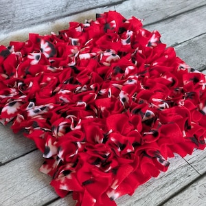 May include: A square, handmade rug with a ruffled texture. The rug is primarily red, with accents of white and black. The fabric appears to be soft and plush, creating a visually appealing and tactile surface.