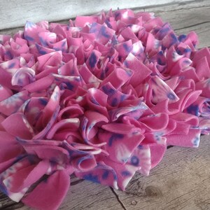 May include: A pink and white fleece dog toy with a tie-dye pattern. The toy is made of strips of fleece tied together to create a textured surface. The fleece is a vibrant pink color with white and blue accents.