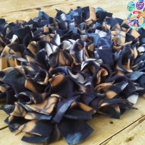 May include: A round, handmade pet mat made of navy blue, tan, and white fleece strips. The mat is designed with a knotted construction, creating a textured surface. The mat is placed on a light brown wooden surface.