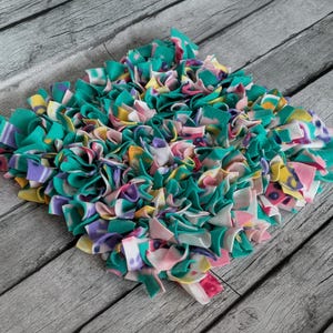 May include: A square, handmade rag rug with a vibrant mix of colors. The rug features teal, pink, purple, yellow, and white fabric strips, creating a textured, decorative piece. The rug is designed for home decor.