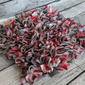 May include: A handmade, square rag rug with a textured surface. The rug is made of fabric strips in shades of red, gray, and white. The rug is placed on a weathered, gray wooden surface.