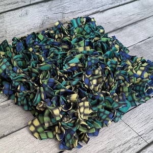 May include: A square, handmade pet mat composed of knotted fleece strips. The mat features a checkered pattern in shades of teal, blue, yellow, and black. The fleece strips are tightly packed, creating a textured surface. The mat is designed for pets.