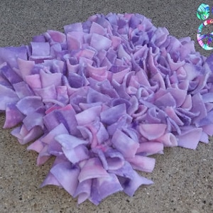 May include: A handmade, square-shaped rug with a textured surface. The rug is made of fabric strips in shades of purple and pink, creating a layered, ruffled effect. The rug is designed for home decor.