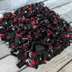 May include: A square, handmade rug composed of black, red, and white fabric strips. The rug has a shaggy texture and is made of multiple layers of fabric. The color scheme is a mix of dark and bright colors.