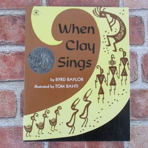 May include: A yellow and brown book cover with the title "When Clay Sings" by Byrd Baylor, illustrated by Tom Bahti. The cover features a stylized Native American design with a flute player and other figures.
