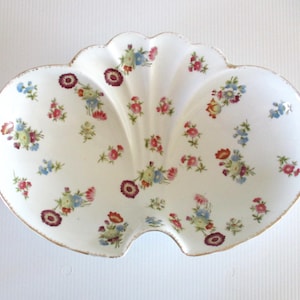 May include: A white ceramic serving dish shaped like a double scallop shell, featuring a delicate floral pattern with small red, pink, blue, and green blossoms scattered across the surface and a thin gold trim along the scalloped edges.