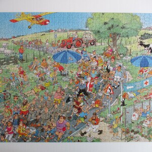 May include: A colorful jigsaw puzzle depicting a busy outdoor marathon scene with numerous runners, spectators, a red tractor, a yellow airplane in the sky, blue umbrellas, and a medical tent with a red cross sign on a green grassy landscape.