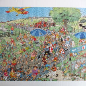 May include: A colorful jigsaw puzzle depicting a crowded outdoor marathon scene with runners on a road, spectators under blue umbrellas, a red tractor, a yellow airplane in the sky, and black and white cows in a green field with a red cross sign.