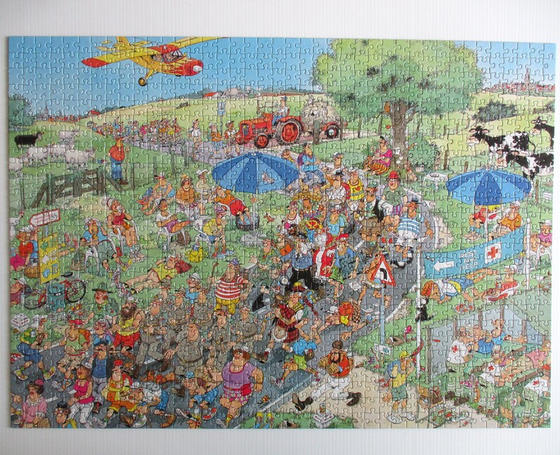 May include: A colorful jigsaw puzzle depicting a crowded outdoor marathon scene with runners on a road, spectators under blue umbrellas, a red tractor, a yellow airplane in the sky, and black and white cows in a green field with a red cross sign.