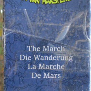 May include: A blue rectangular box with a glossy plastic wrap. The surface features a dark blue pattern and white text that reads: The March, Die Wanderung, La Marche, De Mars. The top edge shows partial yellow text against a dark blue background.