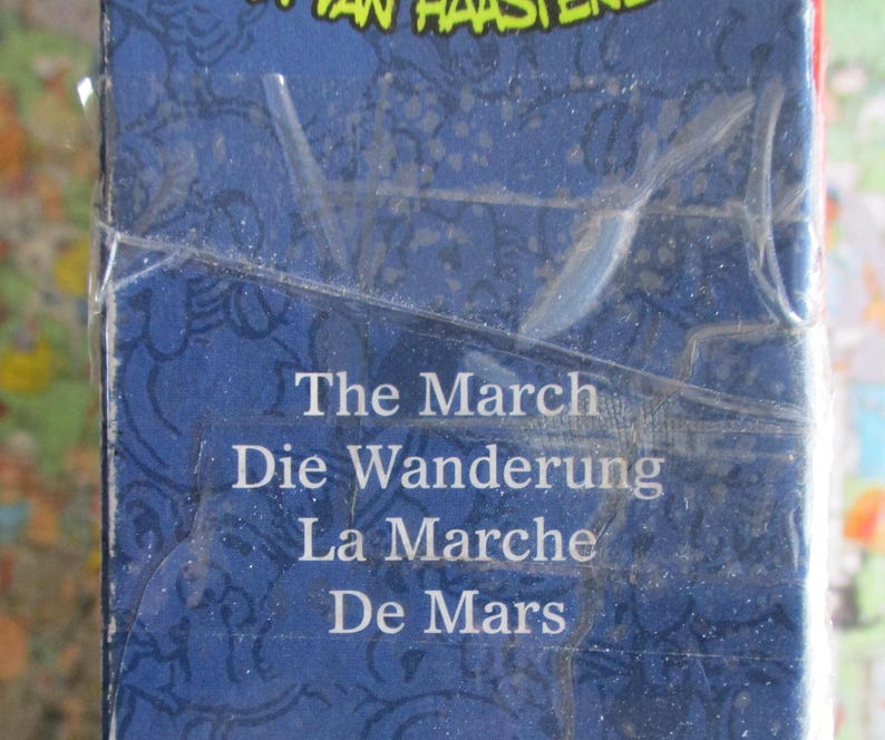 May include: A blue rectangular box with a glossy plastic wrap. The surface features a dark blue pattern and white text that reads: The March, Die Wanderung, La Marche, De Mars. The top edge shows partial yellow text against a dark blue background.