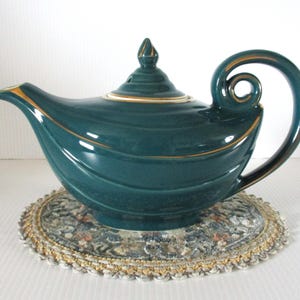 May include: A dark teal ceramic teapot with a glossy finish, featuring a curved handle, a pointed spout, and a matching lid with a finial. The teapot is decorated with subtle gold-colored trim along the edges, handle, and lid, showcasing a smooth, rounded body design.