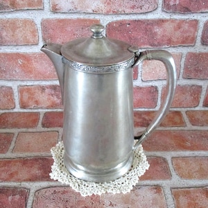 May include: A vintage silver-colored coffee pot with a lid and handle, resting on a cream-colored crocheted doily. The pot has a decorative band around the top and is set against a red brick background.