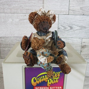 May include: A brown and tan teddy bear figurine wearing a blue and white dress and a red bow. The bear is holding a smaller brown and tan teddy bear figurine wearing a blue and white dress with a floral pattern. The figurines are sitting on a white box with the text "Computer pal SCREEN SITTER" printed on it.