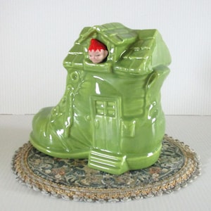 May include: A whimsical, green ceramic boot-shaped house with a small figure in a red hat peeking out of the roof. The boot has a door and window details. It sits on a decorative, round mat.