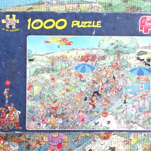 May include: A 1000-piece jigsaw puzzle box by Jan van Haasteren featuring a colorful, chaotic cartoon illustration of a crowded outdoor event with a red tractor, a yellow airplane, blue umbrellas, and numerous people in a rural landscape.