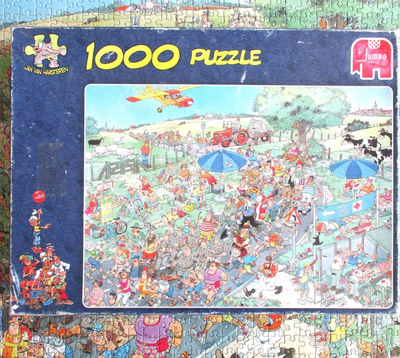 May include: A 1000-piece jigsaw puzzle box by Jan van Haasteren featuring a colorful, chaotic cartoon illustration of a crowded outdoor event with a red tractor, a yellow airplane, blue umbrellas, and numerous people in a rural landscape.