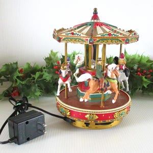 May include: A miniature carousel with a red and green roof, gold accents, and multiple painted horses. The base is red with gold trim and features an on-off switch and a power port. A black power adapter with a cord is connected to the carousel.