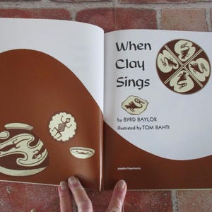 May include: An open book titled "When Clay Sings" by Byrd Baylor, illustrated by Tom Bahti. The cover features a brown background with a white bird design and the title in black text.