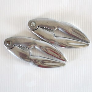 May include: Two shiny, silver-colored crab crackers. Each cracker has a claw-like design with a toothed edge for gripping and cracking shells. The tools are made of metal and are designed for seafood preparation.