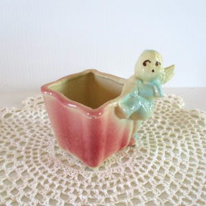 May include: A square ceramic planter featuring a glossy pink and cream gradient finish. A small, light blue and cream-colored cherub figure with wings is attached to the side of the planter, appearing to climb over the rim of the container.