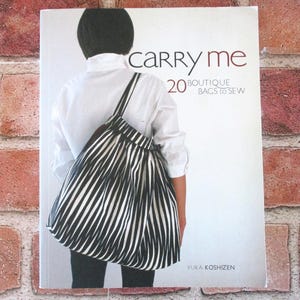 May include: A softcover book titled Carry Me: 20 Boutique Bags to Sew by Yuka Koshizen. The cover features a person wearing a white shirt and carrying a black and white striped fabric bag with a gathered top and thin shoulder straps.
