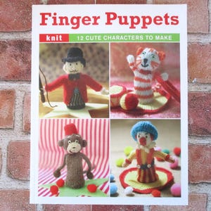 May include: A softcover book titled Finger Puppets: 12 Cute Characters to Make. The cover features four photographs of knitted finger puppets, including a ringmaster, a striped cat, a monkey with a red fez, and a clown, all in vibrant colors.
