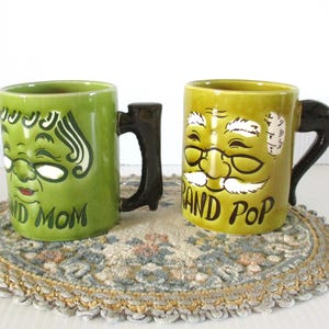 May include: Two ceramic mugs with black handles. One is green with a face design and the words "GRAND MOM". The other is yellow with a face design and the words "GRAND POP". Both mugs are on a decorative doily.