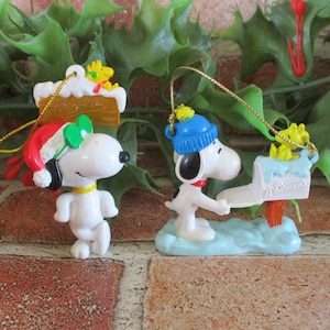 May include: Two Snoopy Christmas ornaments. One Snoopy wears a Santa hat and the other wears a blue hat and delivers mail. Both ornaments have a yellow string for hanging. The ornaments are white with black accents and are on a green background.