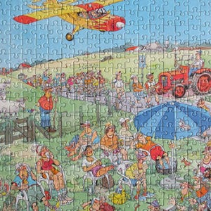 May include: A jigsaw puzzle featuring a cartoon illustration of a rural scene with a yellow airplane flying overhead, a red tractor on a path, a group of people walking, and a crowd of spectators sitting under a large blue umbrella in a field.