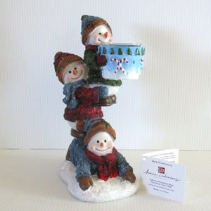 May include: A whimsical snowman figurine with three snowmen stacked on top of each other. The snowmen wear hats, scarves, and sweaters in shades of blue, red, and brown. The top snowman holds a blue and white container with candy cane designs.