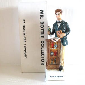 May include: A ceramic figurine of a man in a dark green jacket and tan pants, holding a small figurine and a book labeled "TAB." The figurine stands next to a cabinet displaying miniature bottles. The box reads "MR. BOTTLE COLLECTOR" and "BY TRADER-TAB COMPANY."