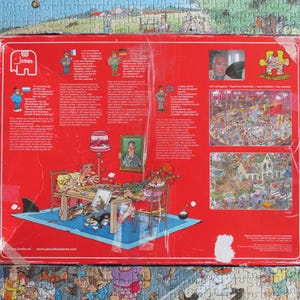 May include: A rectangular red cardboard jigsaw puzzle box featuring a cartoon illustration of two people assembling a puzzle at a table. The box includes the Jumbo logo, text in multiple languages, a portrait of Jan van Haasteren, and two smaller images of completed puzzles.