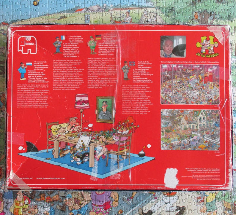May include: A rectangular red cardboard jigsaw puzzle box featuring a cartoon illustration of two people assembling a puzzle at a table. The box includes the Jumbo logo, text in multiple languages, a portrait of Jan van Haasteren, and two smaller images of completed puzzles.