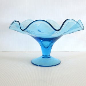 May include: A light blue glass pedestal bowl with a ruffled rim and a dark blue rim. The bowl has a flared, wavy edge and a pedestal base. The bowl is made of glass and is likely vintage.