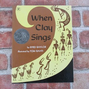 May include: A children's book cover with a yellow and brown background. The title is "When Clay Sings" by Byrd Baylor, illustrated by Tom Bahti. The cover features a silver coin with a Native American design.