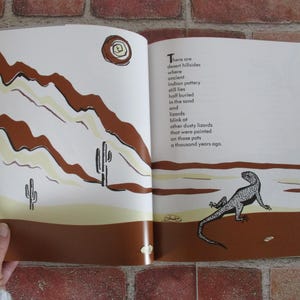 May include: An open book with a brown and white illustration of a desert landscape with cacti and a lizard on the right page. The left page has text that reads "There are desert hillsides where ancient Indian pottery still lies half buried in the sand and lizards blink at other dusty lizards that were painted on those pots a thousand years ago."