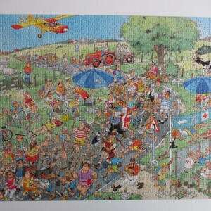 May include: A jigsaw puzzle featuring a colorful, busy cartoon illustration of a crowded outdoor event. The scene includes a yellow airplane flying overhead, a red tractor, blue umbrellas, and numerous people walking along a road.